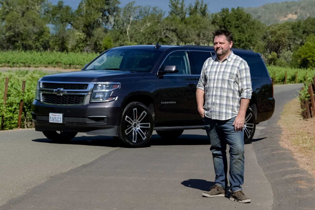 Ryan Olson stands on a lane between vineyards with his SUV behind him